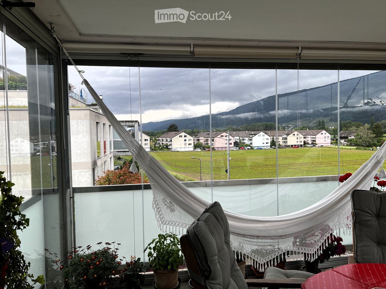 Balcony with large windows offering a scenic view of a green field, mountains, and buildings. Features a hanging chair, a small table, potted plants, and a hammock.