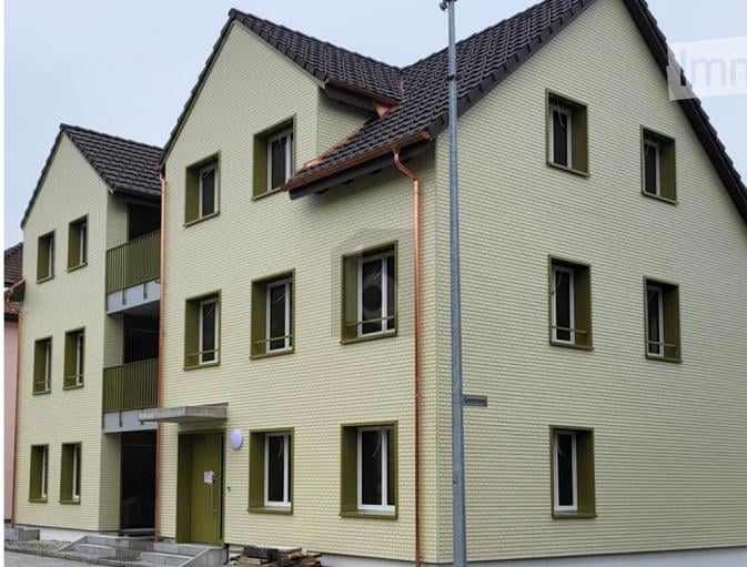 Two-story residential building with light yellow siding, green shutters, and multiple windows. Features a small balcony on the upper floor and a covered entrance area.