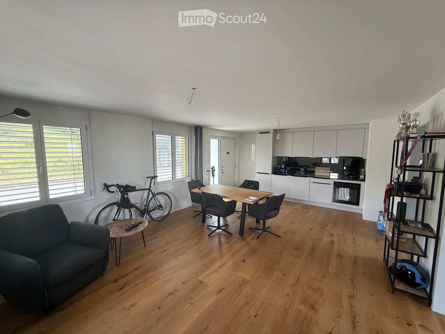 Open-plan living and dining area with wooden flooring, white walls, and large windows. Features a sofa, coffee table, dining table with chairs, bicycle, shelf with trophies, and a modern kitchen with white cabinets and stainless steel appliances.