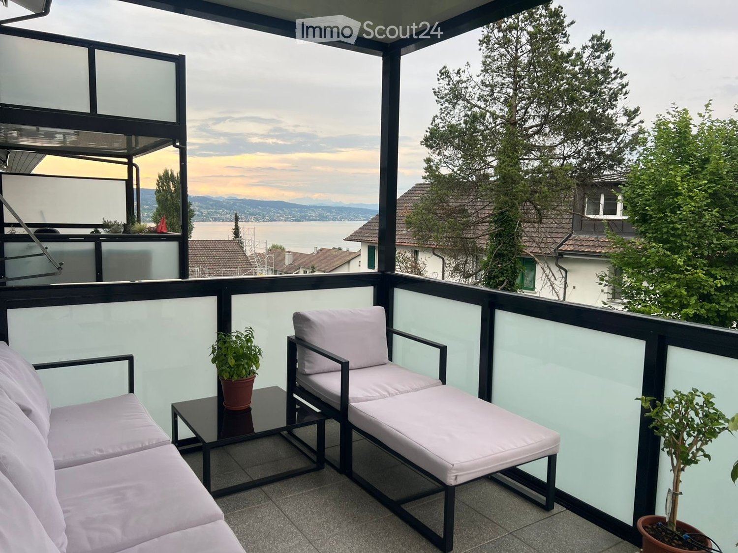 Balcony with a view of the lake and mountains, featuring two white lounge chairs, a small black table, and potted plants.