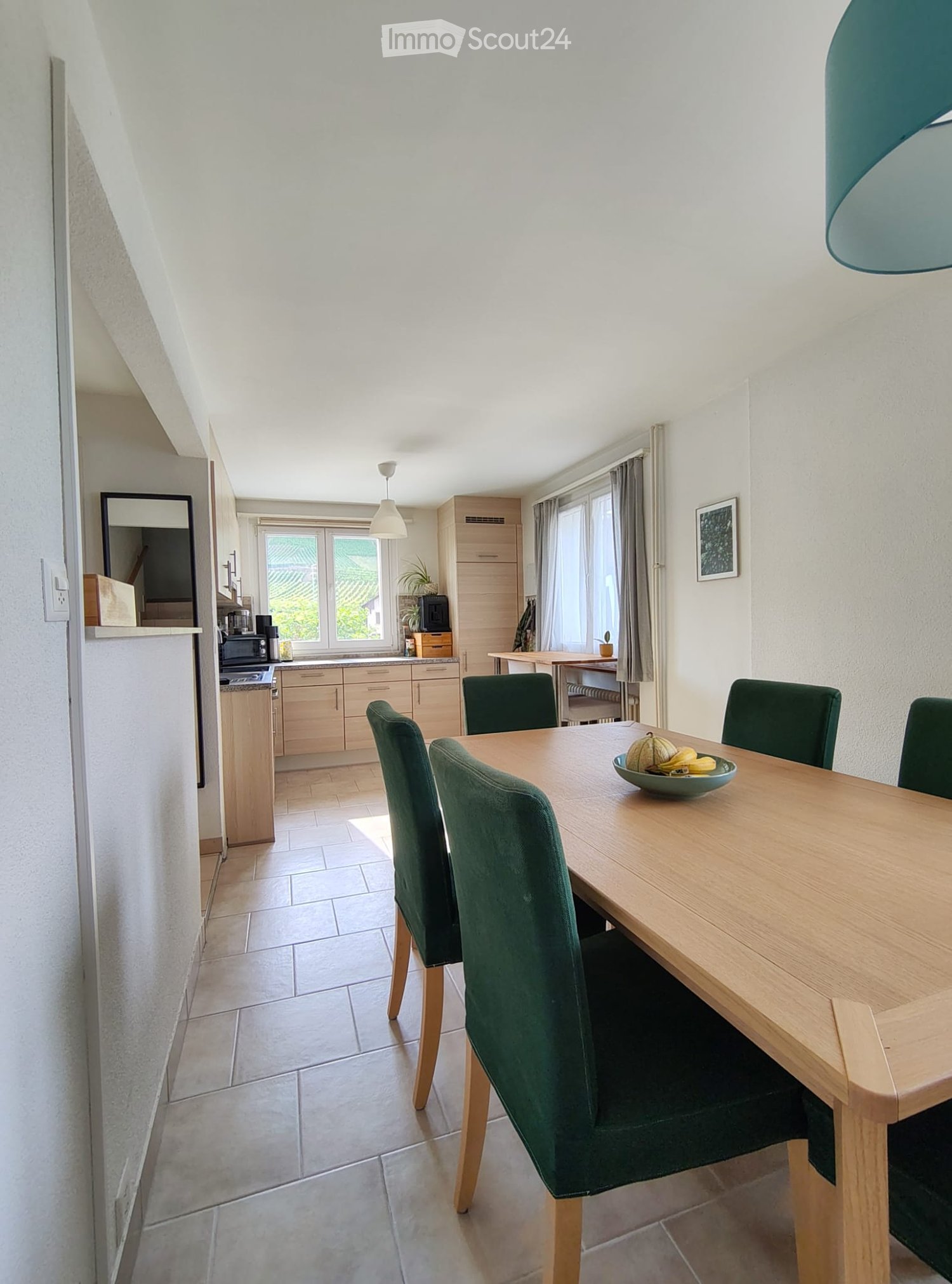 Bright kitchen and dining area with a wooden dining table, green chairs, and a bowl of fruit on the table. The kitchen features light wooden cabinets, a microwave, and a refrigerator.