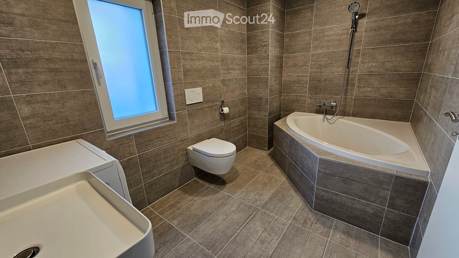 Modern bathroom with grey tiled walls and floor, featuring a corner bathtub, toilet, and sink.