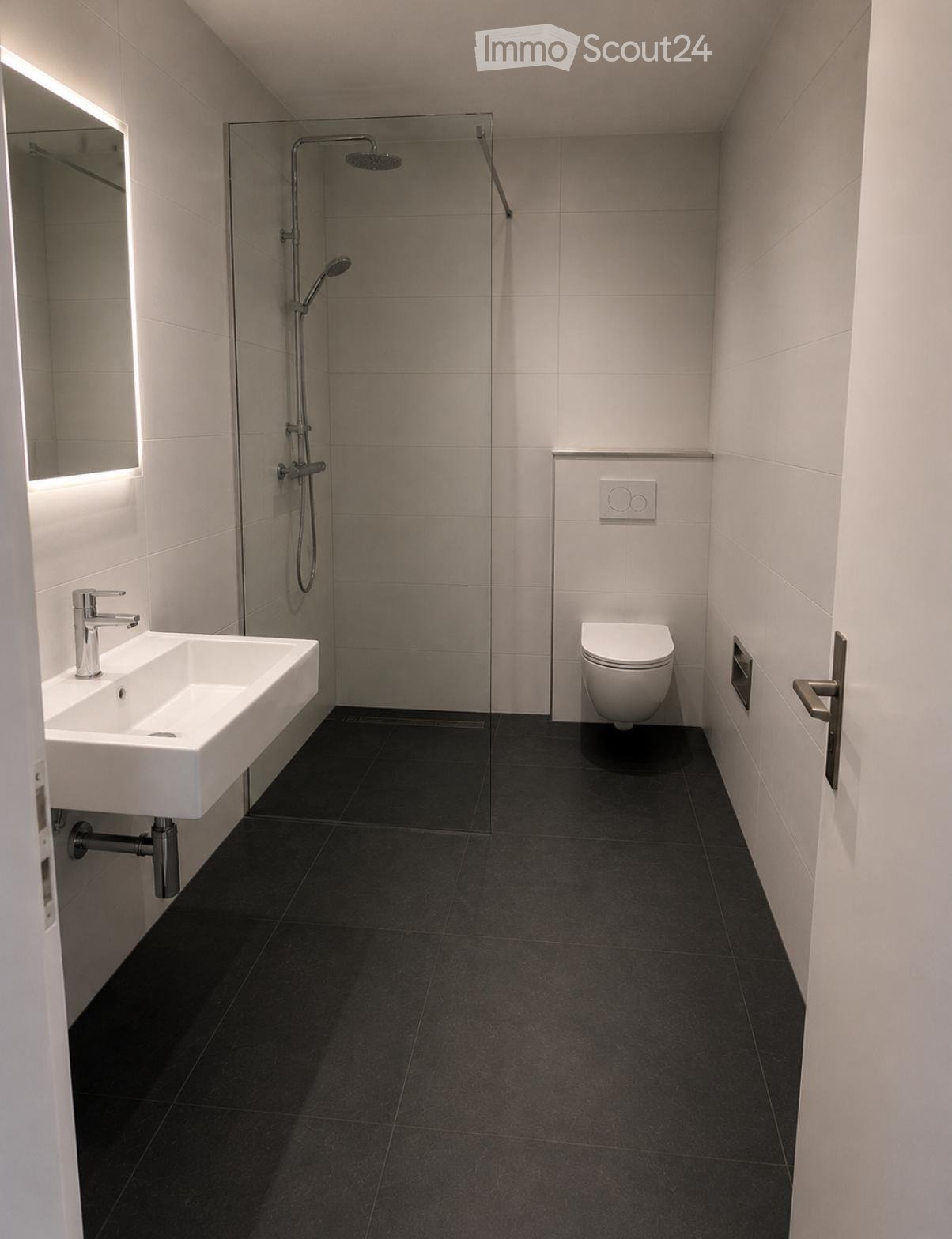Modern bathroom with a white sink, toilet, and glass-enclosed shower. Features dark tiled flooring and light grey tiled walls.