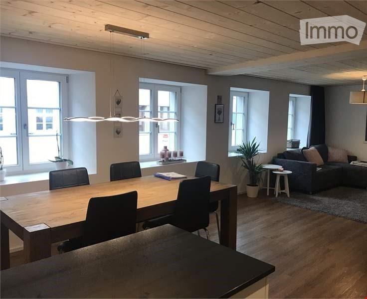 Open-plan living and dining area with wooden flooring, large windows, a dining table with chairs, a sofa, a small stool, and potted plants.