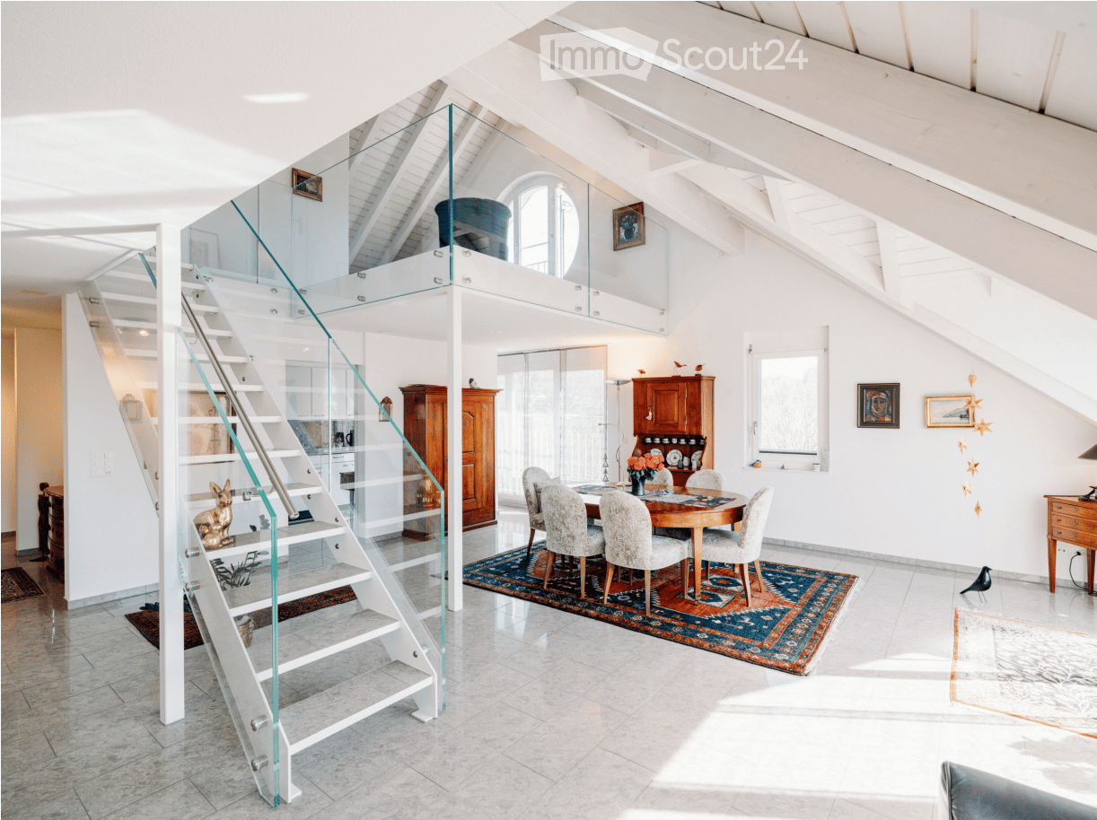 Modern, open-concept living and dining area with high ceilings, exposed wooden beams, and a glass staircase leading to an upper level. The dining area features a round wooden table with six upholstered chairs, a colorful area rug, and a vase with flowers