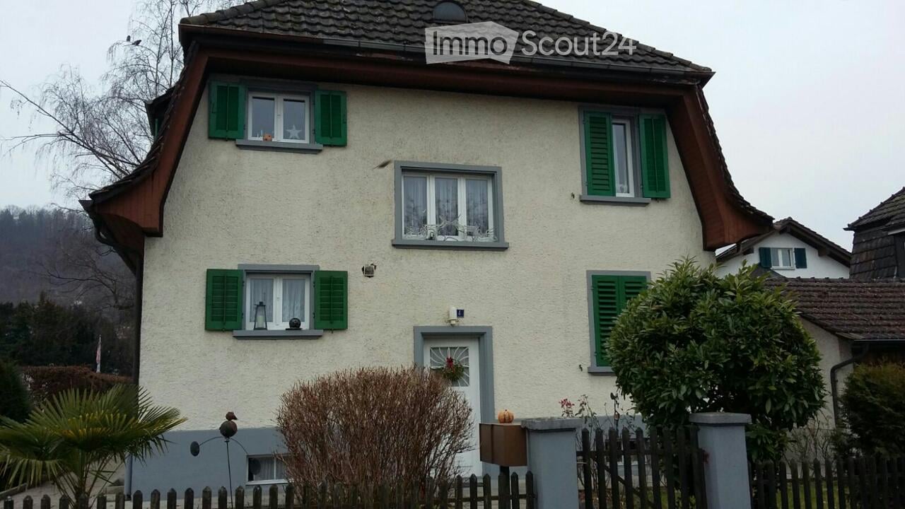 Two-story house with white siding, green shutters, and multiple windows. Features a small garden with a wooden fence and decorative items.
