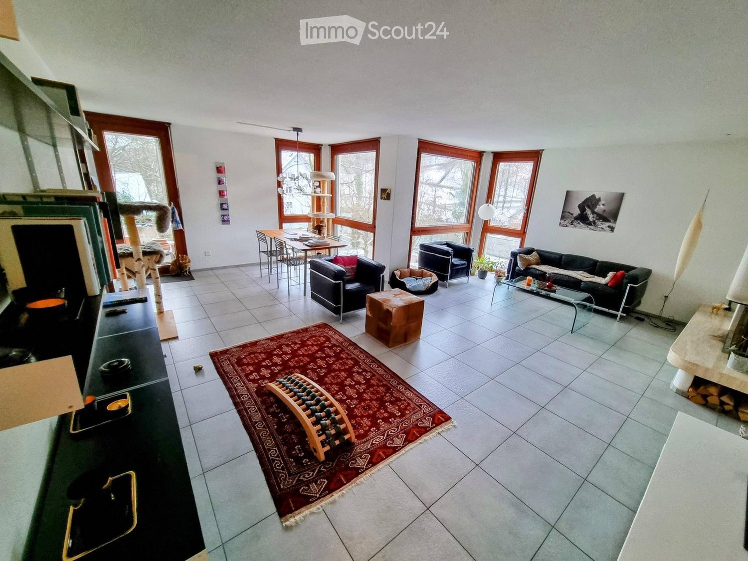 Bright living room with large windows, tiled floor, red rug, sofa, coffee table, cat tree, dining table, and chairs.