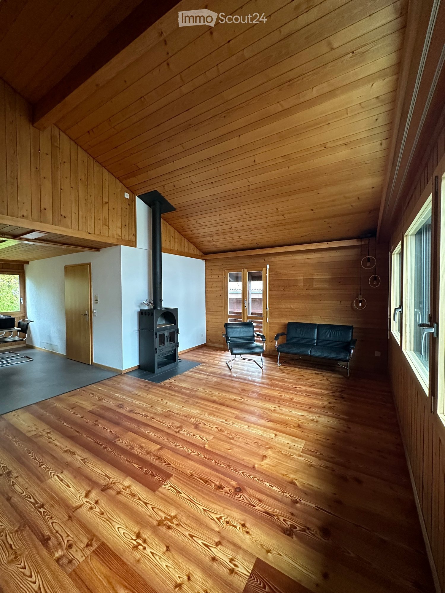 Spacious living room with wooden floors, exposed wooden ceiling beams, and a central fireplace. Features a dark green sofa and matching armchair, with large windows and a door leading outside.