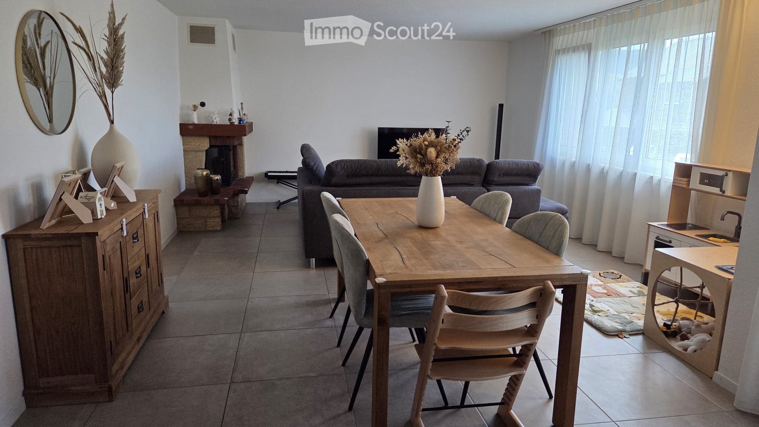 Bright living and dining area with a grey sofa, wooden dining table, fireplace, and decorative vases.