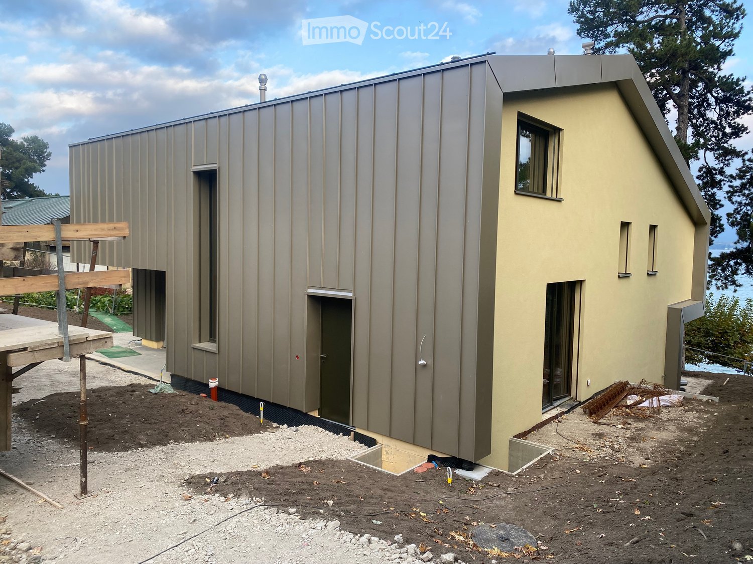 House in construction, grey metal and beige facade, 2 main entrances, windows, and outdoor space in front