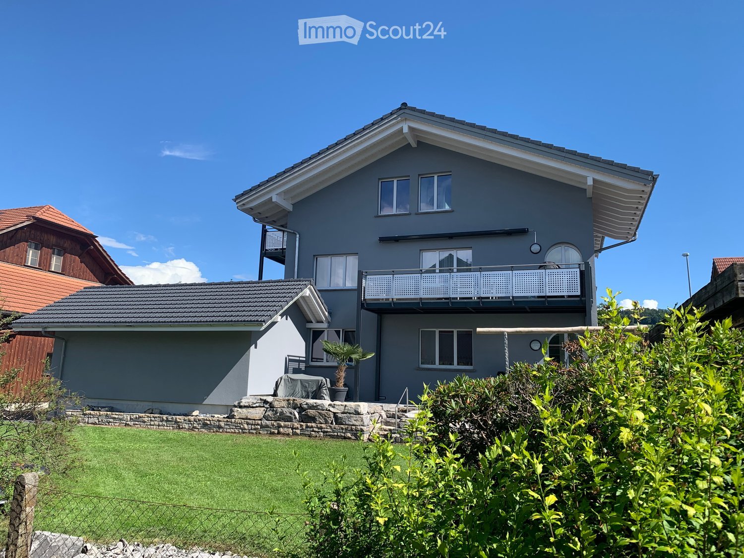 Two-story modern house with grey exterior, white trim, multiple windows, and a balcony on the upper level. The house features a well-maintained lawn and surrounding greenery.