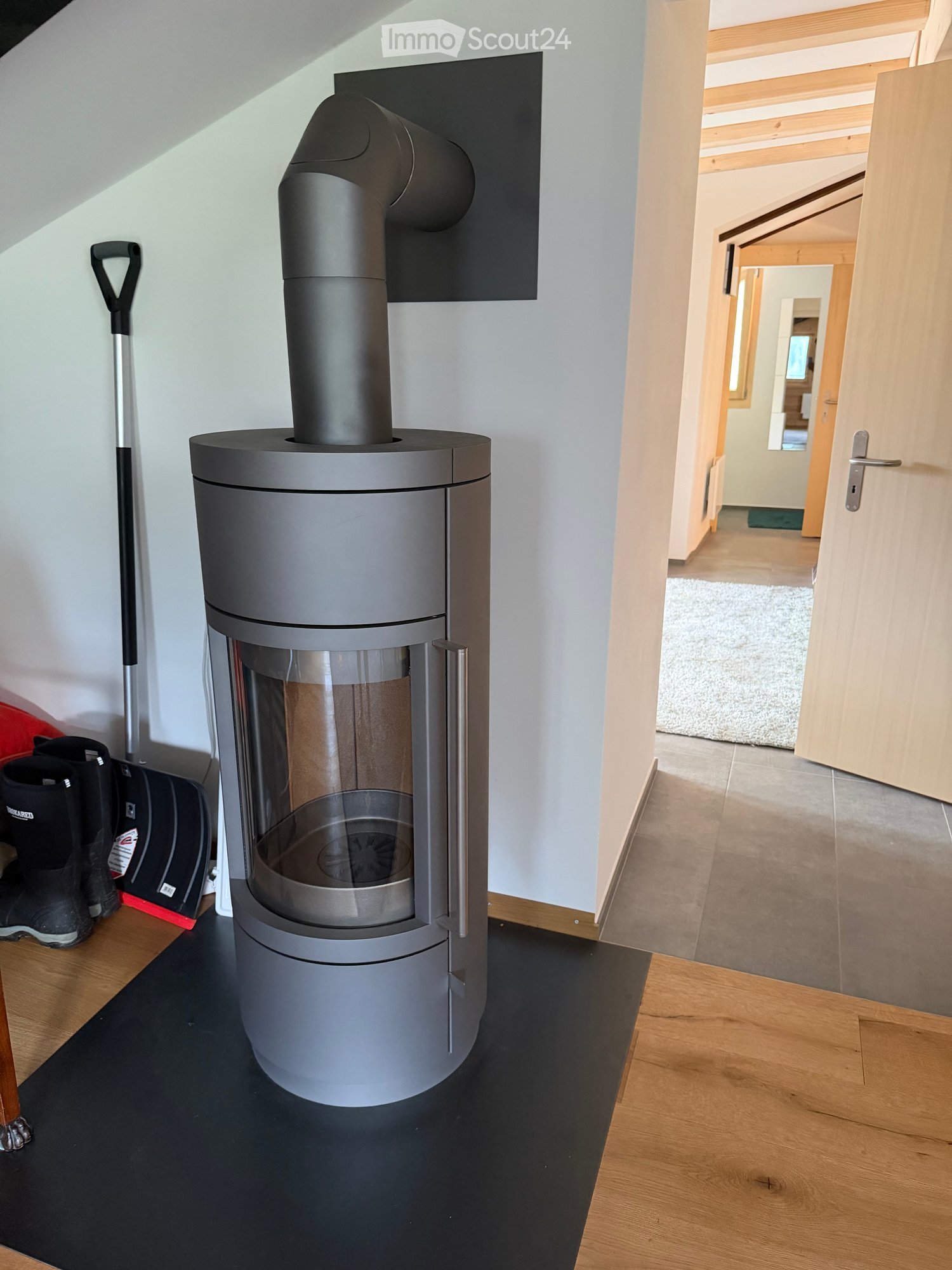 Hallway with wooden flooring, open doorway leading to another room, modern grey wood-burning stove with chimney, and a few items like boots and a broom leaning against the wall.