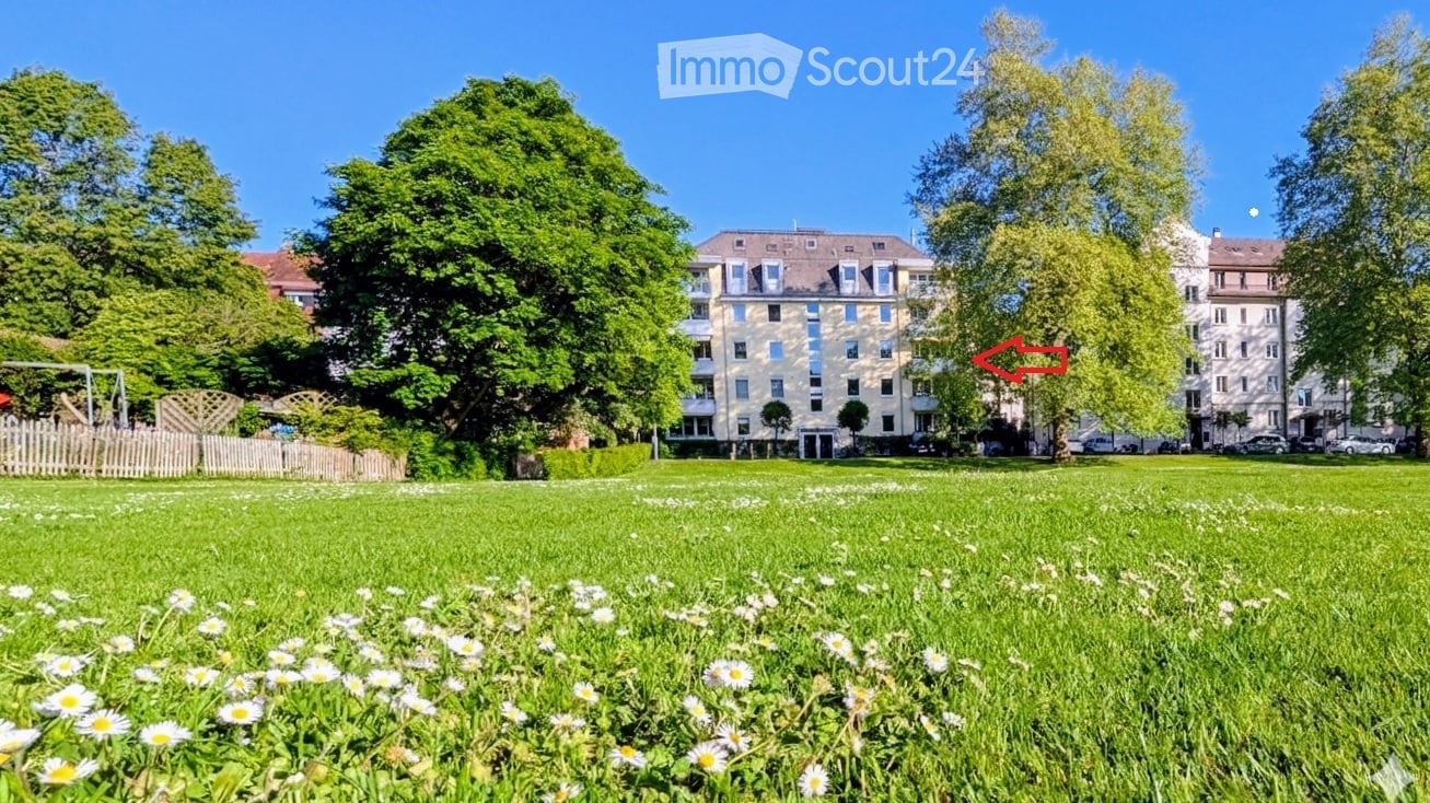 A well-maintained park area with green grass, white daisies, and trees, with a multi-story residential building in the background.