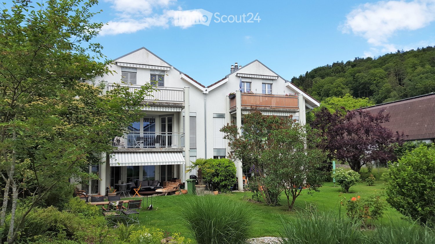 A two-story white house with balconies, surrounded by a well-maintained garden with outdoor furniture and greenery.