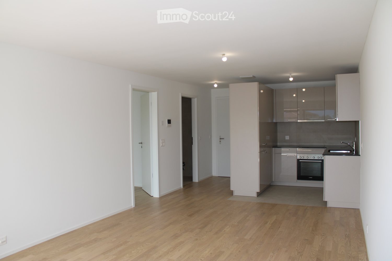 Empty room with light wooden flooring, white walls, and a small kitchen area featuring built-in cabinets and appliances.