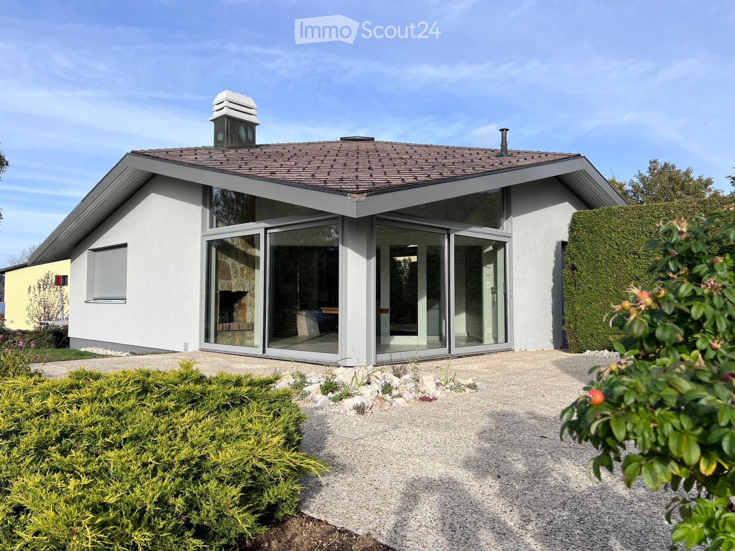 Modern single-story house with white exterior, large glass sliding doors, and a tiled roof. The house features a raised ground floor and is surrounded by neatly trimmed hedges and gravel pathways.