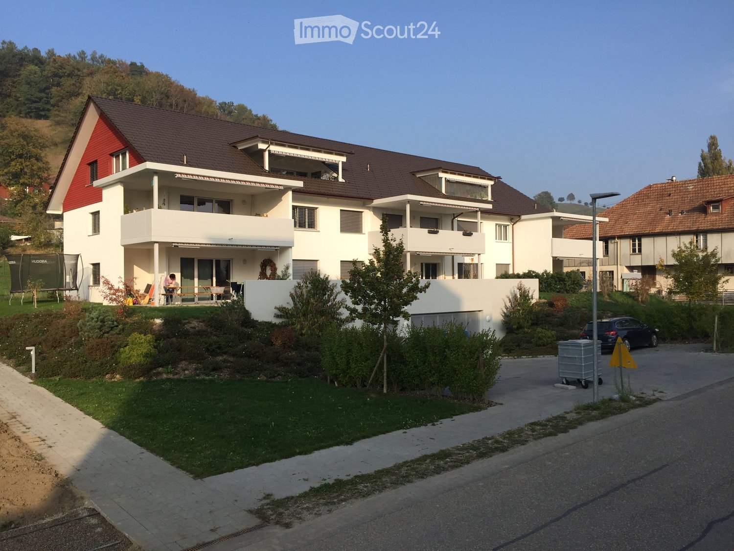 Two-story residential building with white walls, red roof accents, and balconies. The surrounding area includes a lawn, trees, a parked car, and a small playground with trampolines.