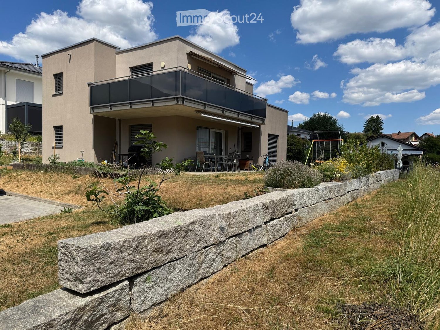 Modern two-story house with a balcony, patio seating area, and garden with stone wall and plants.