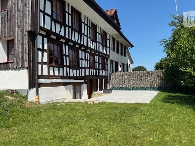 Traditional half-timbered house with white plaster walls, wooden beams, and multiple windows. The house has a sloped roof and is surrounded by a well-maintained lawn and a stone wall.
