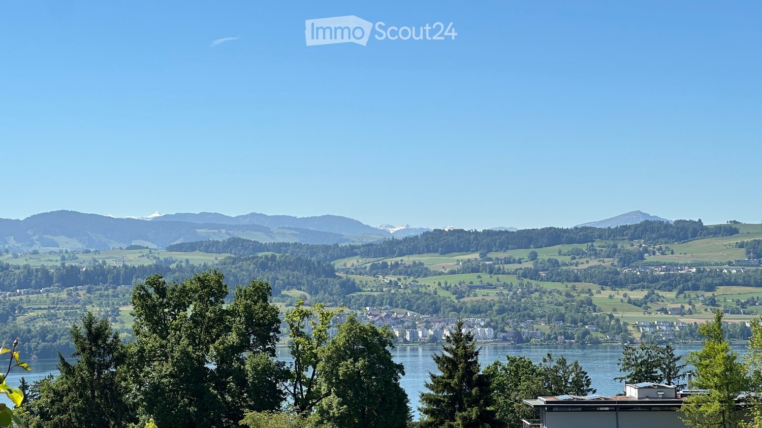 Panoramic view of a lake surrounded by lush green hills and distant mountains under a clear blue sky.