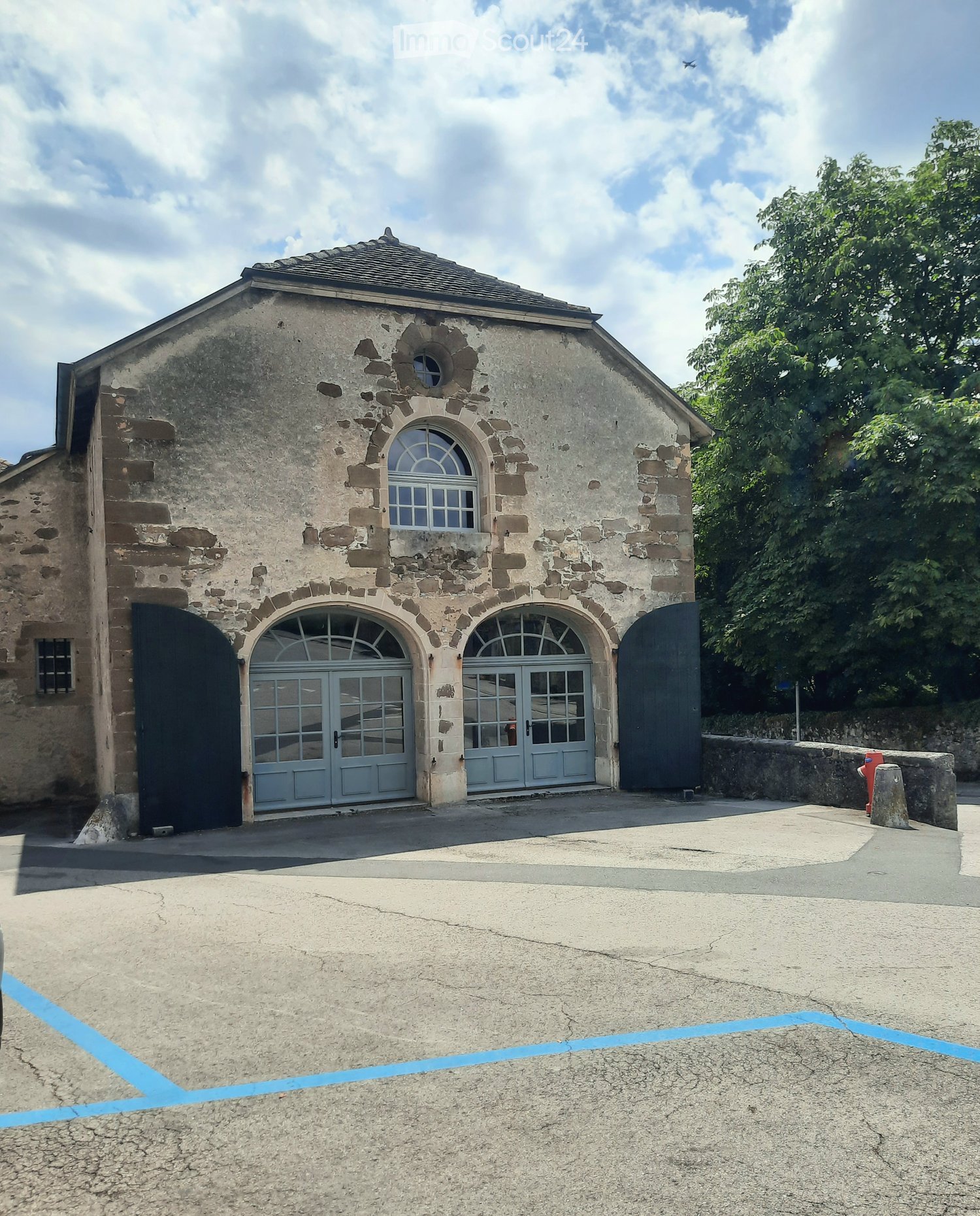 Stone building with arched doors and windows, surrounded by greenery and a paved area.