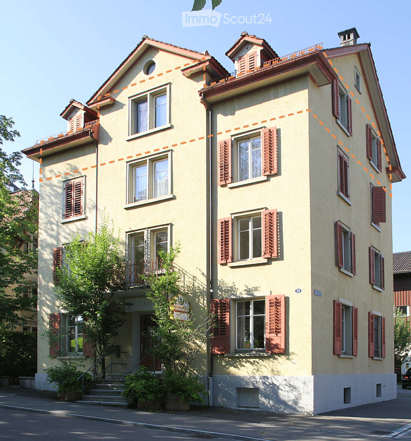 A multi-story residential house with light yellow walls, red shutters, and multiple windows. Features include a balcony on the second floor, a small garden area, and a staircase leading to the entrance.