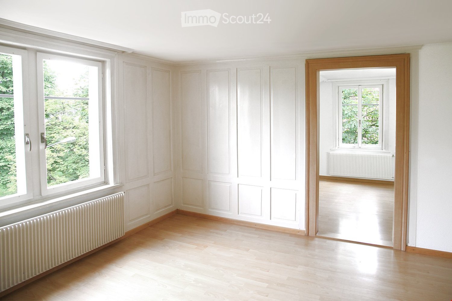 Empty room with light wooden flooring, white walls, large windows, and radiators. No furniture or equipment present.