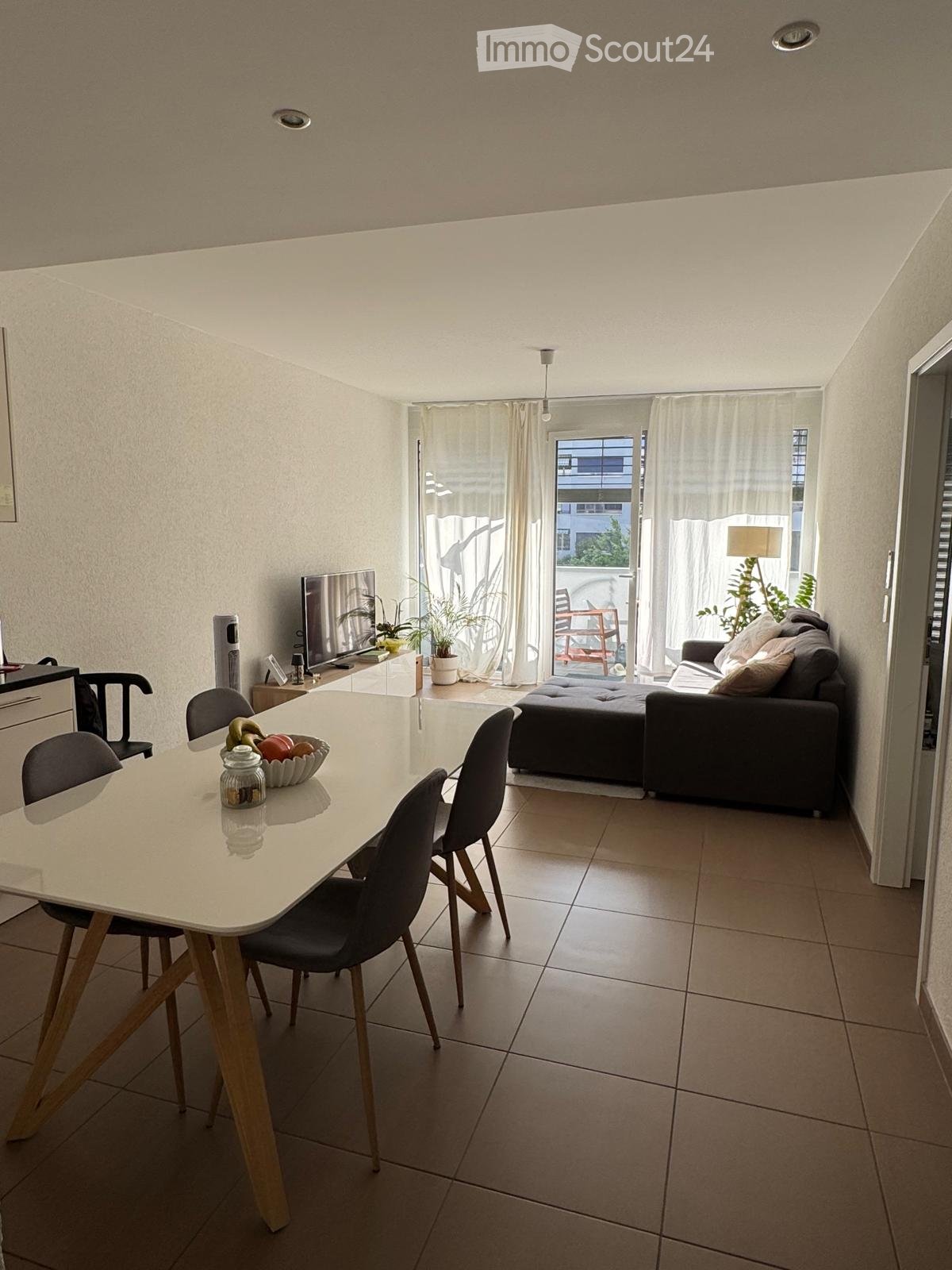 Open-plan living and dining area with a sofa, ottoman, dining table, chairs, and a TV. Large windows with curtains lead to a balcony with outdoor furniture.