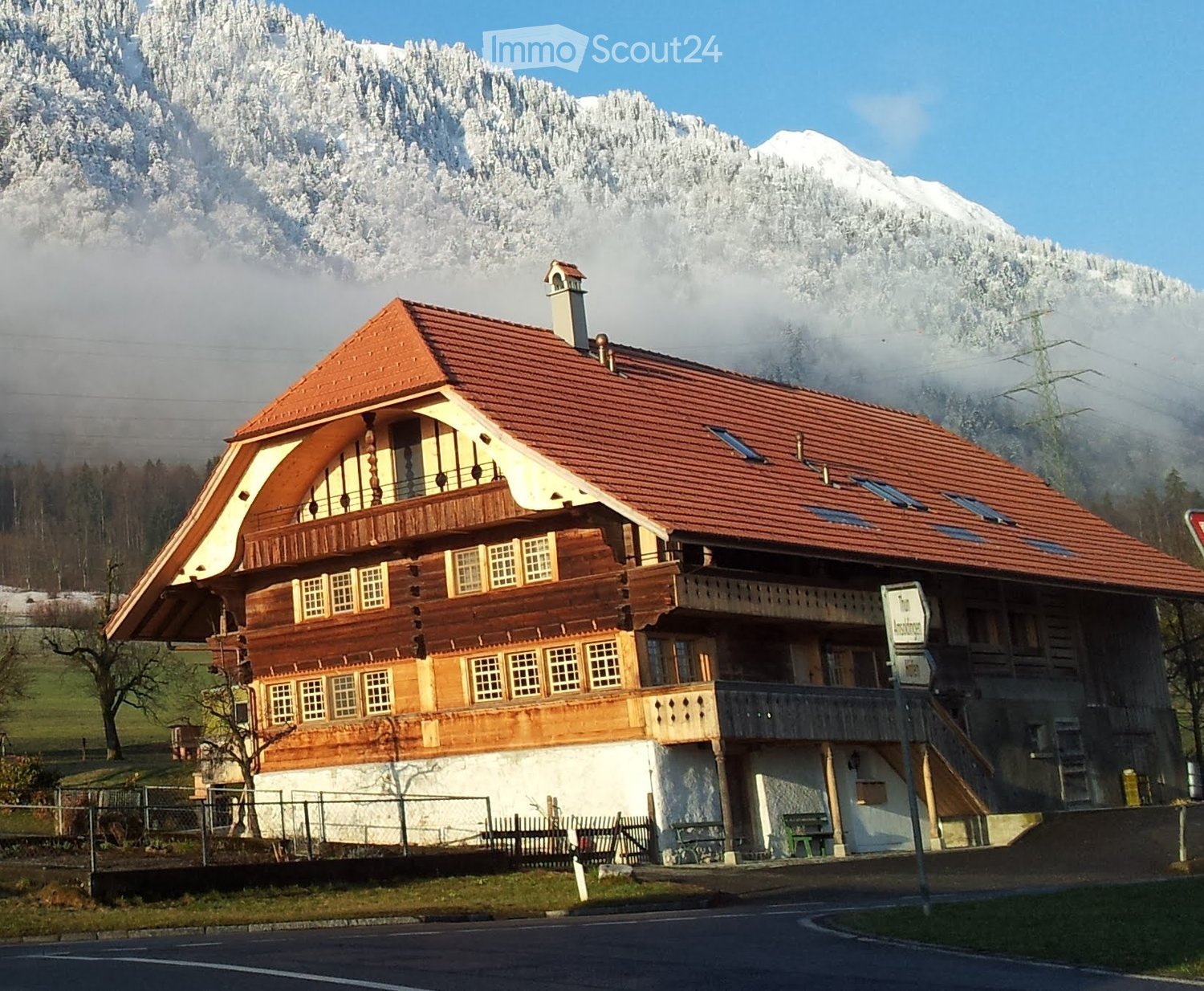 A two-story wooden house with a red-tiled roof, large windows, and a balcony overlooking a snowy mountain landscape.