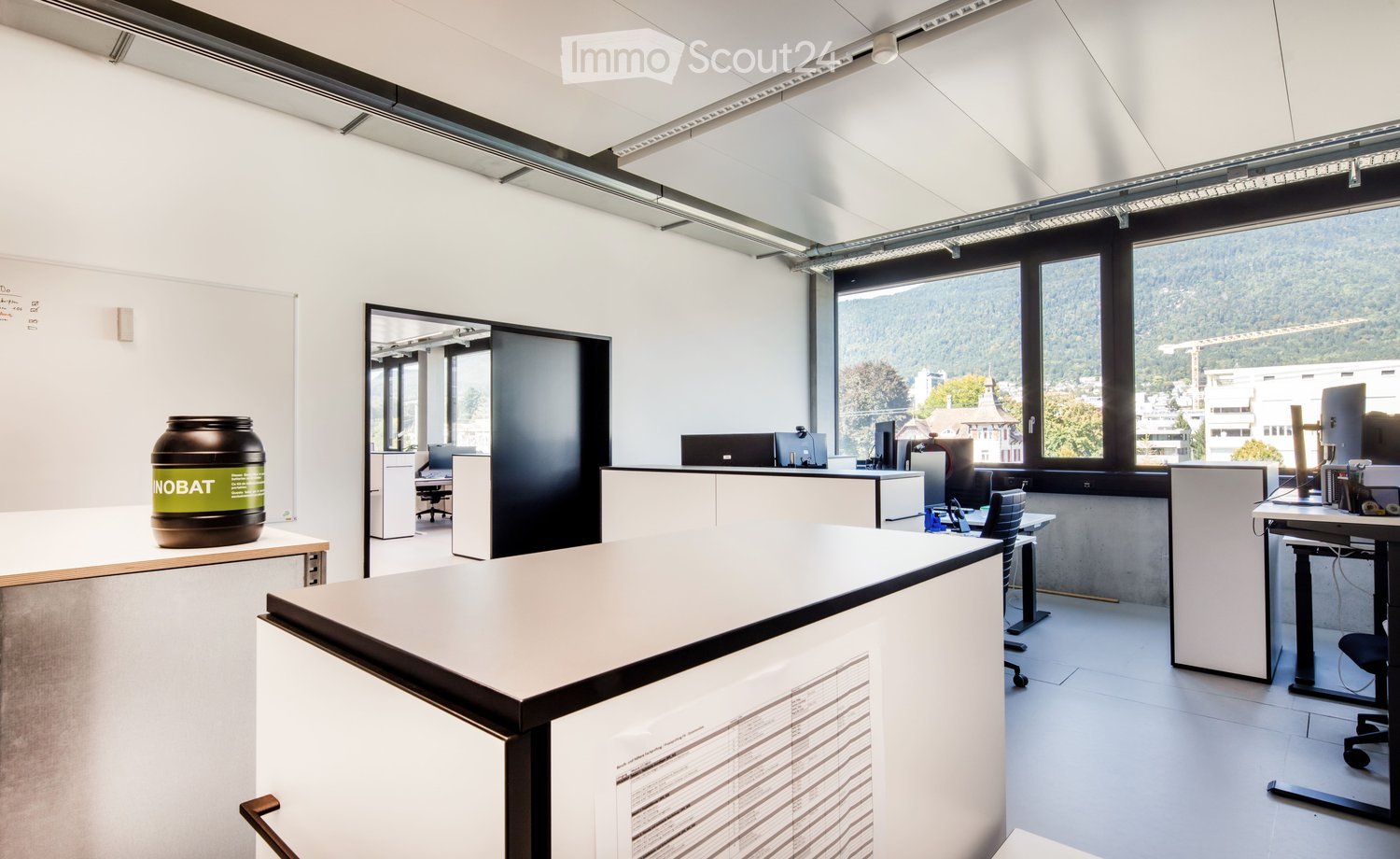 Modern office space with white walls, large windows, desks, chairs, and a large screen. A large container is placed on a table near the window.