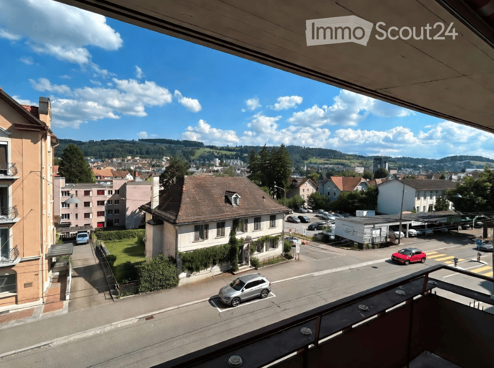 Balcony overlooking a town with houses, parked cars, and a scenic landscape with hills and trees.