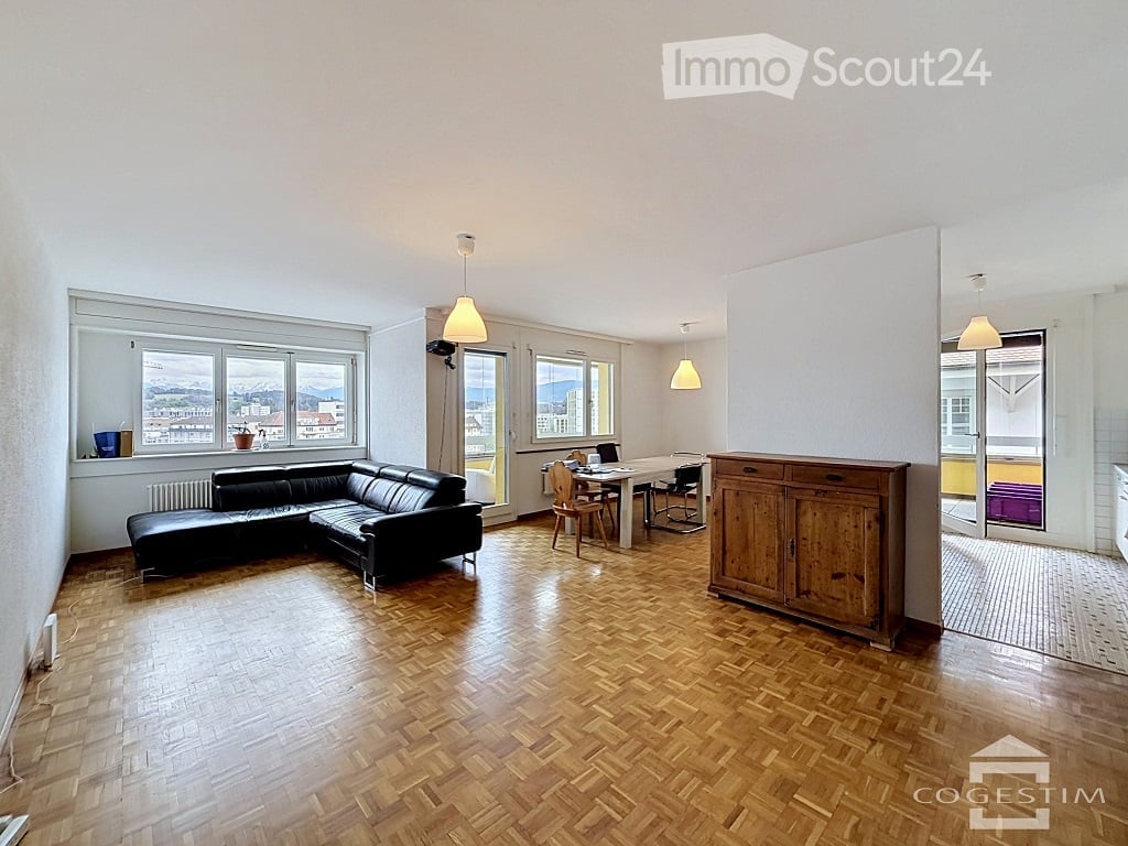 Open-plan living and dining area with wooden flooring, large windows offering a scenic view, a black leather sofa, a dining table with chairs, and a wooden cabinet.