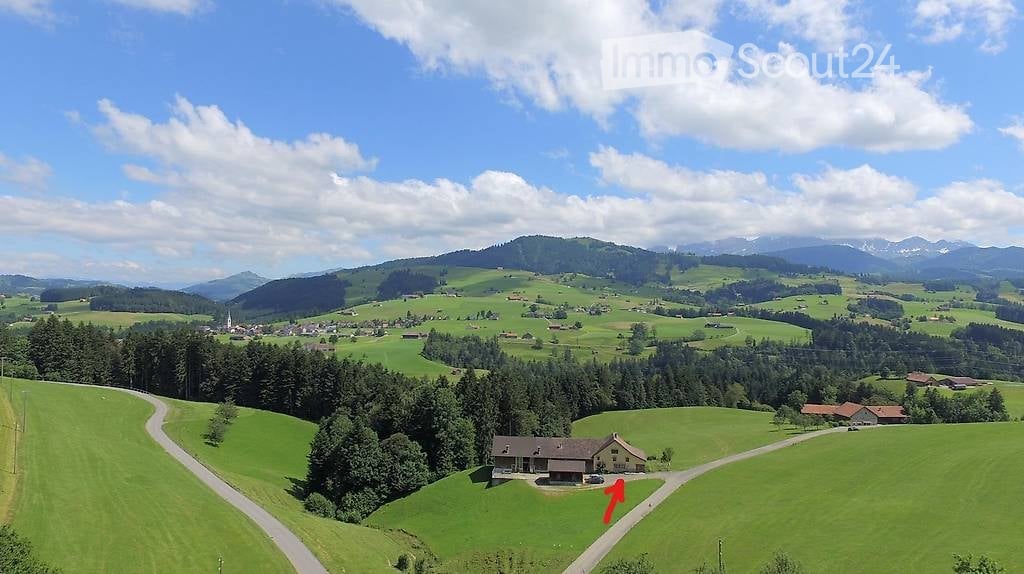 A panoramic view of a lush green valley with rolling hills, scattered trees, and a small village in the distance. A house is visible on a hill, surrounded by open fields and a winding road.