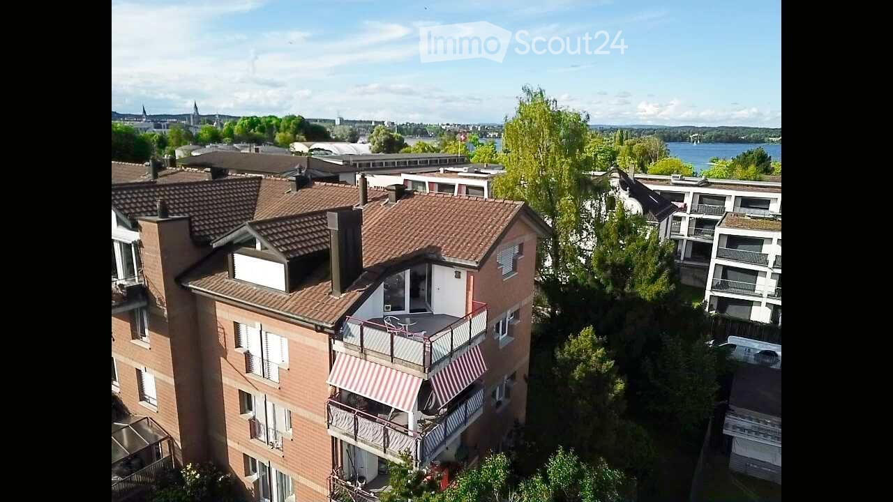Aerial view of a multi-story residential building with balconies, surrounded by trees and a scenic view of a lake and cityscape in the background.