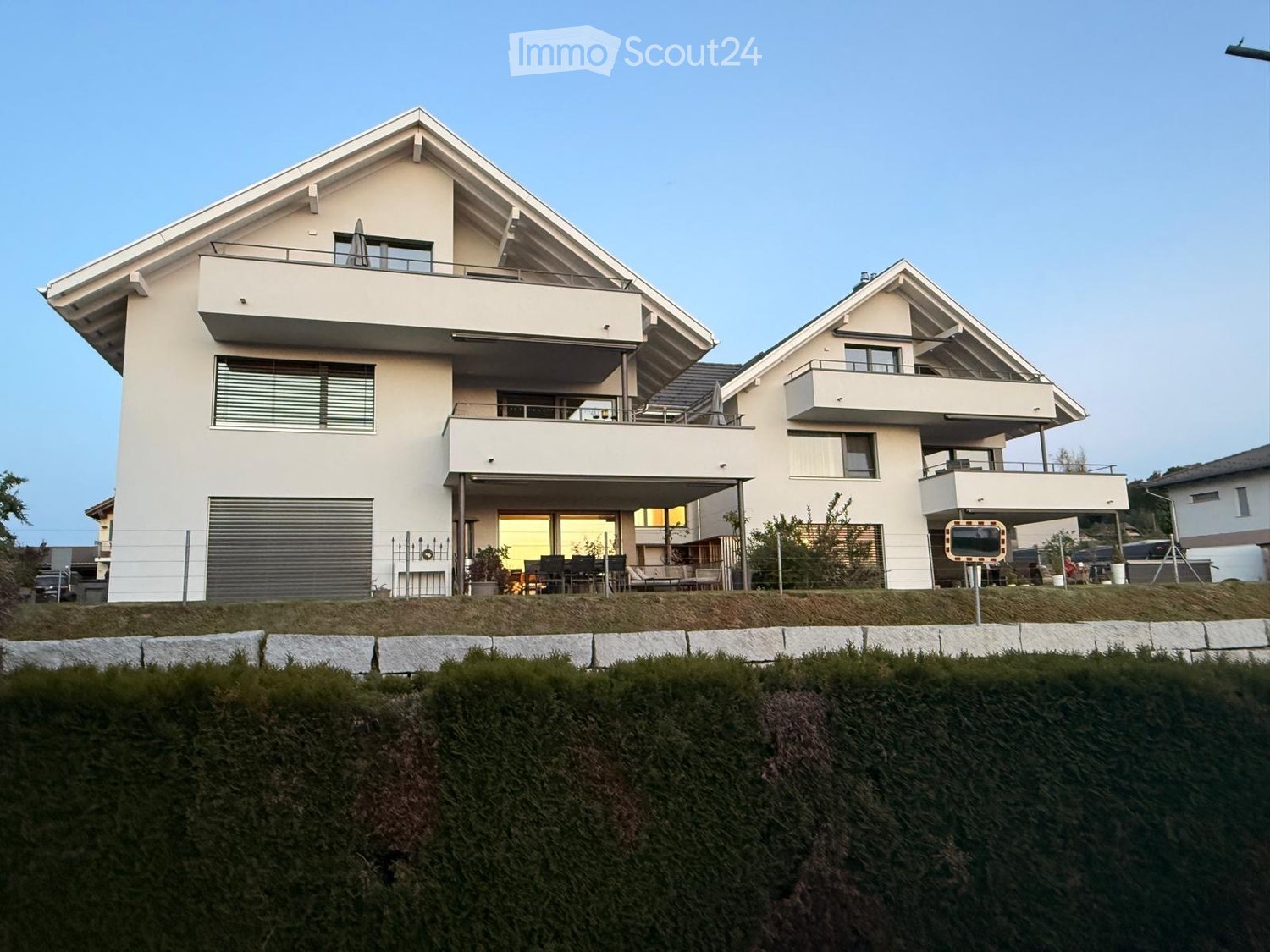 Modern two-story house with white exterior, large windows, balconies, and a garage. The house is situated on a raised ground floor with a well-maintained lawn and hedge in front.