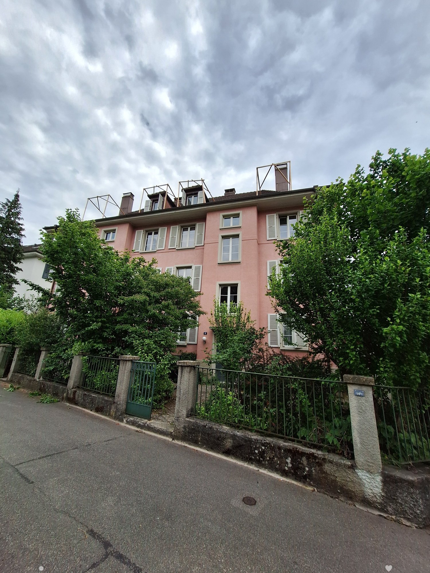 Pink two-story house with multiple windows, shutters, and a small rooftop terrace, surrounded by greenery and a metal fence.