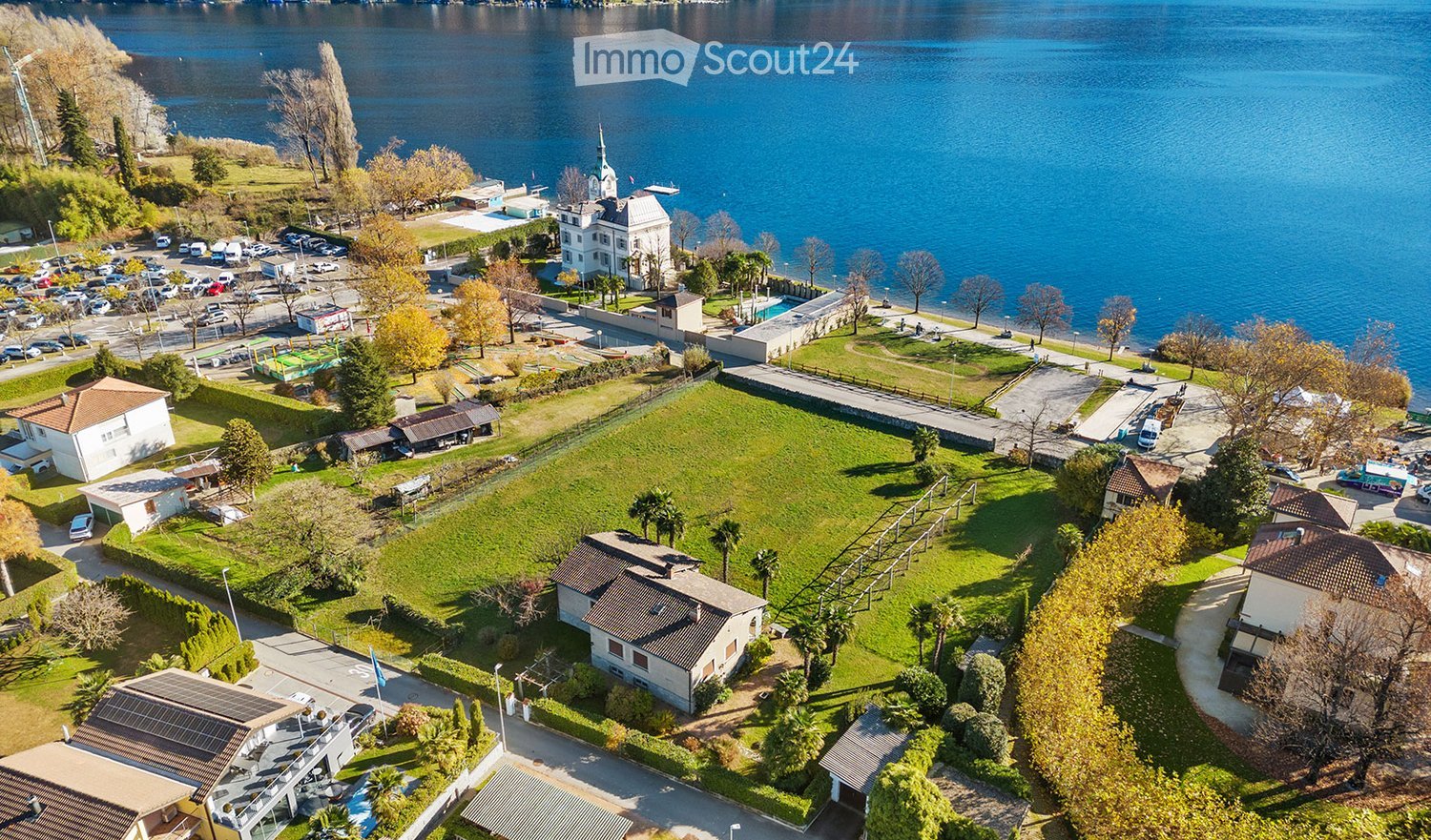 Aerial view of a lakeside area with a large grassy field, several buildings, parking lot, and a view of the lake.