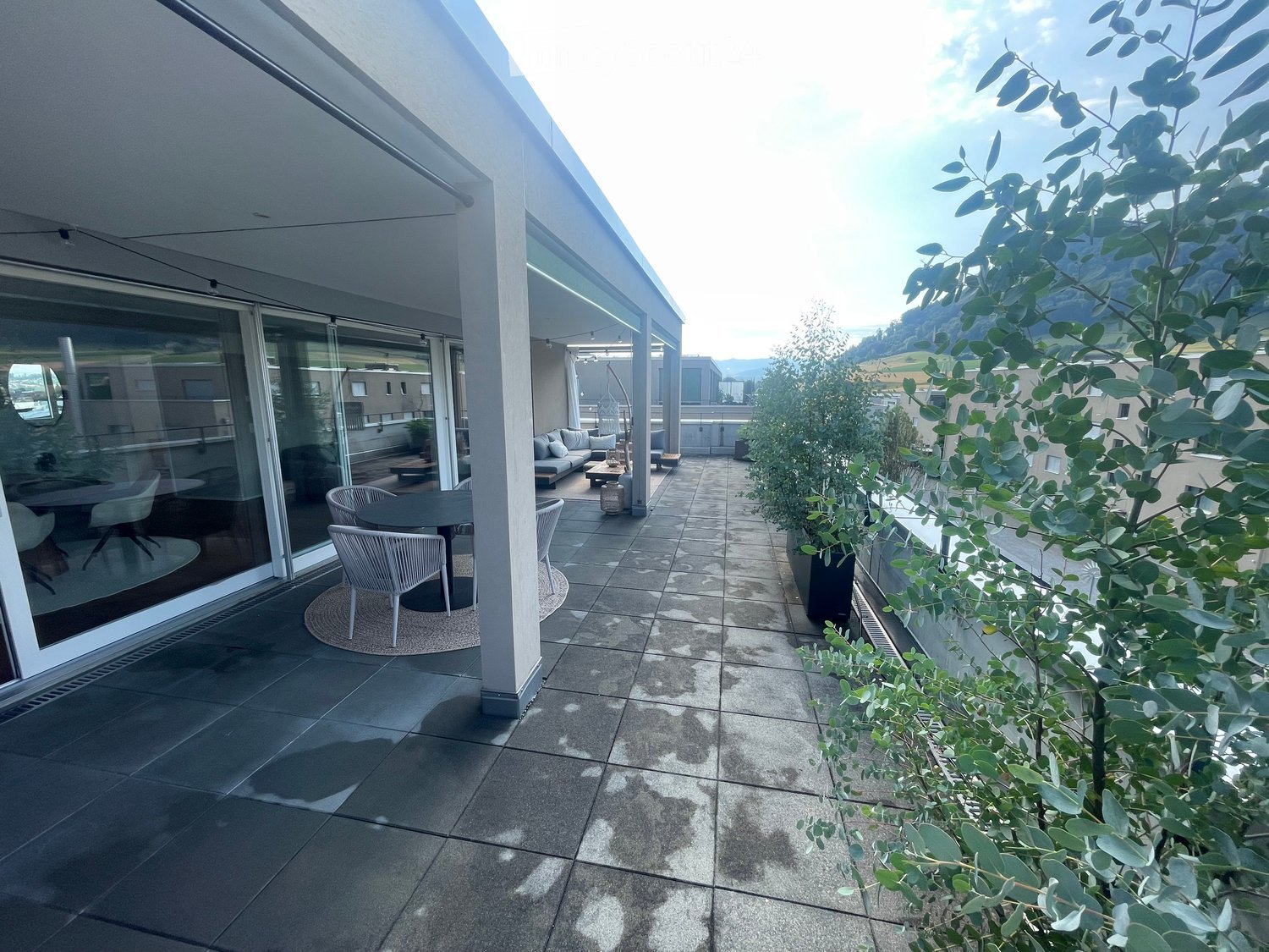 Modern balcony with tiled floor, outdoor furniture including a table and chairs, large glass sliding doors leading to indoor space, and lush green plants.