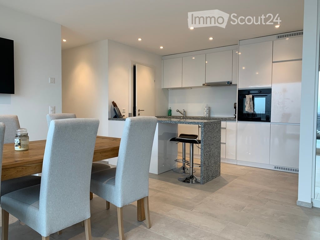 A modern, open-plan kitchen and dining area with white cabinets, a wooden dining table, grey chairs, and a granite countertop. The kitchen features a stove, sink, and built-in oven, with a small bar stool at the counter.