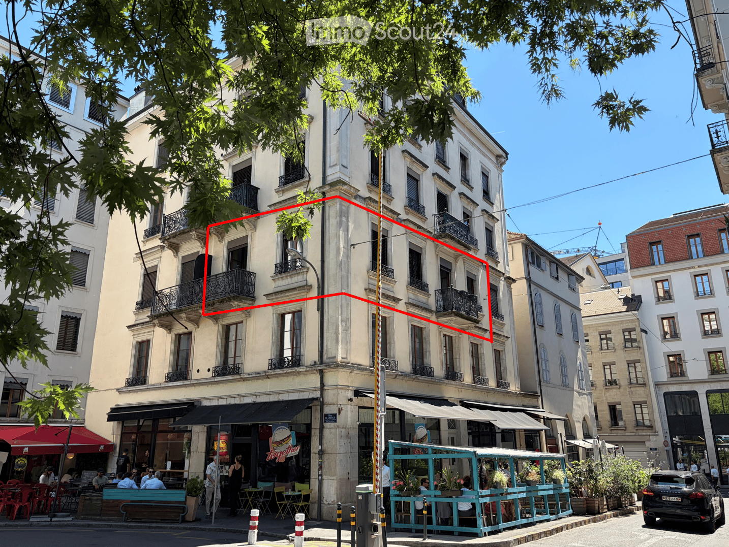 A multi-story building with classic architecture, featuring balconies and large windows. The ground floor has a restaurant with outdoor seating, and the street is lined with trees and other buildings.