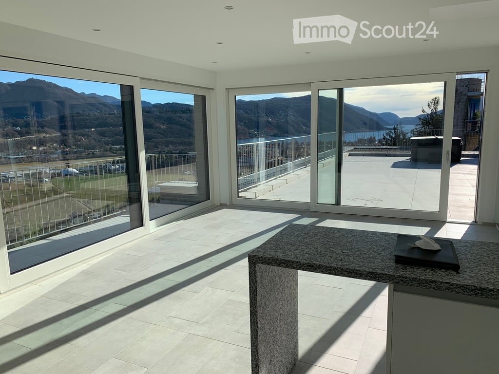 Bright, modern room with large glass sliding doors leading to a balcony, offering scenic mountain and lake views. The room features a polished tile floor and a central stone table.