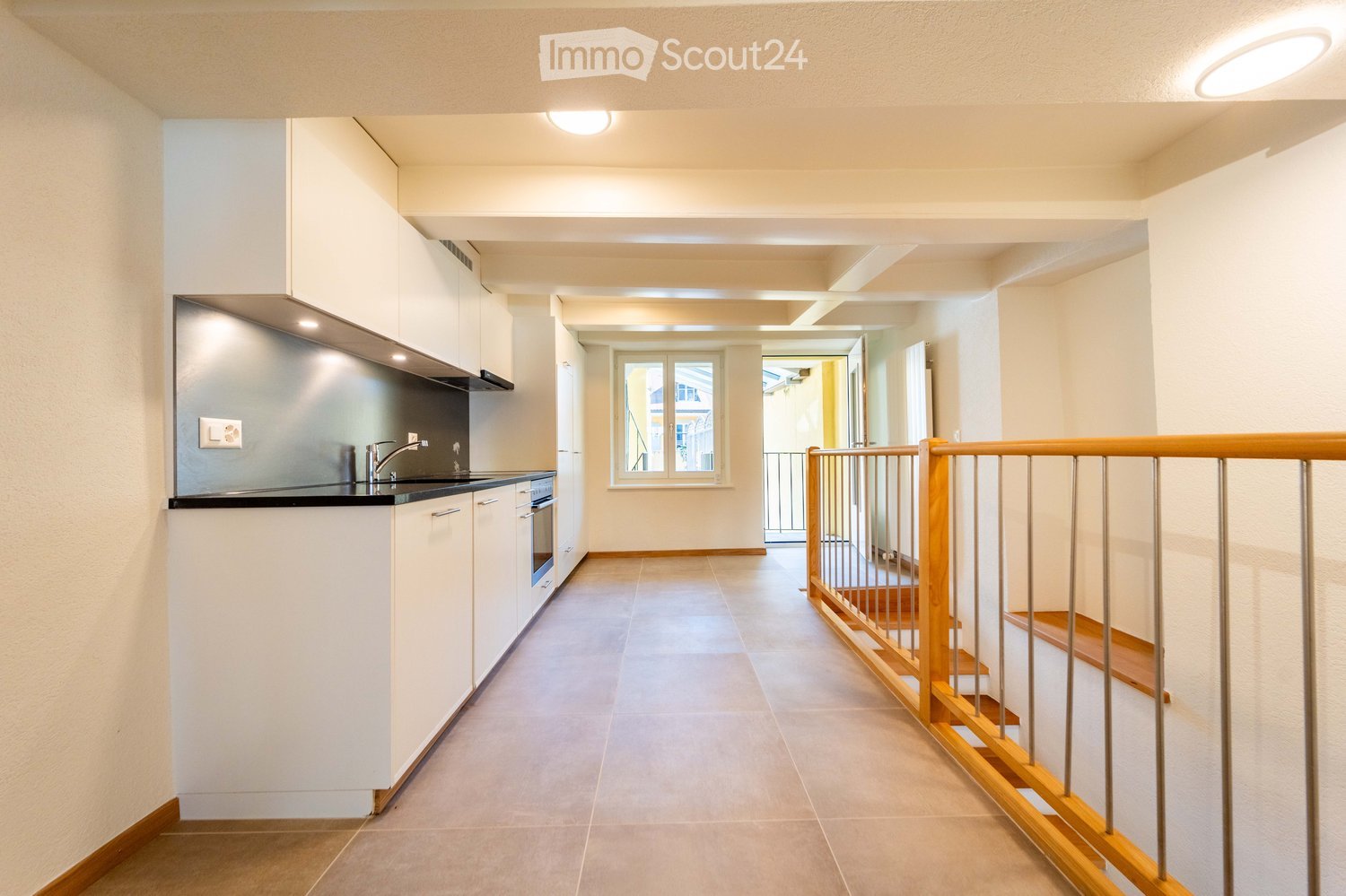 Modern hallway with tiled floor, white walls, built-in kitchen area on the left, staircase with wooden railing leading upwards, and a window at the end.