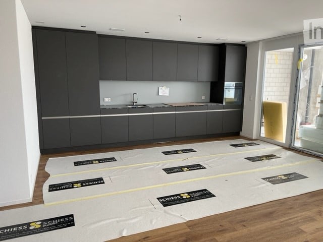 Modern kitchen with dark grey cabinets, a sink, and a wooden floor. Protective plastic sheets are laid out on the floor, likely for renovation or installation purposes.