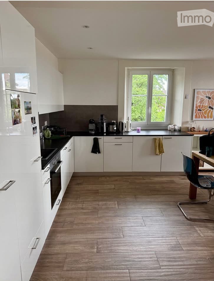 Modern kitchen with white cabinets, black countertops, and wooden flooring. Features include a refrigerator, oven, coffee maker, and a small dining area with a chair and table.