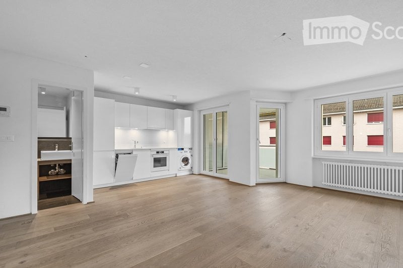 Completely empty room with wooden flooring, white walls, and modern white kitchen cabinets visible in the background. No furniture or equipment present.