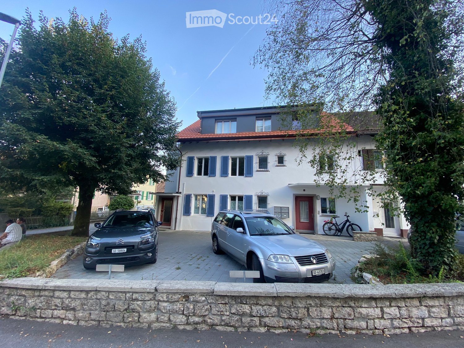 Two-story house with white walls, blue shutters, red roof, and a driveway with two parked cars.