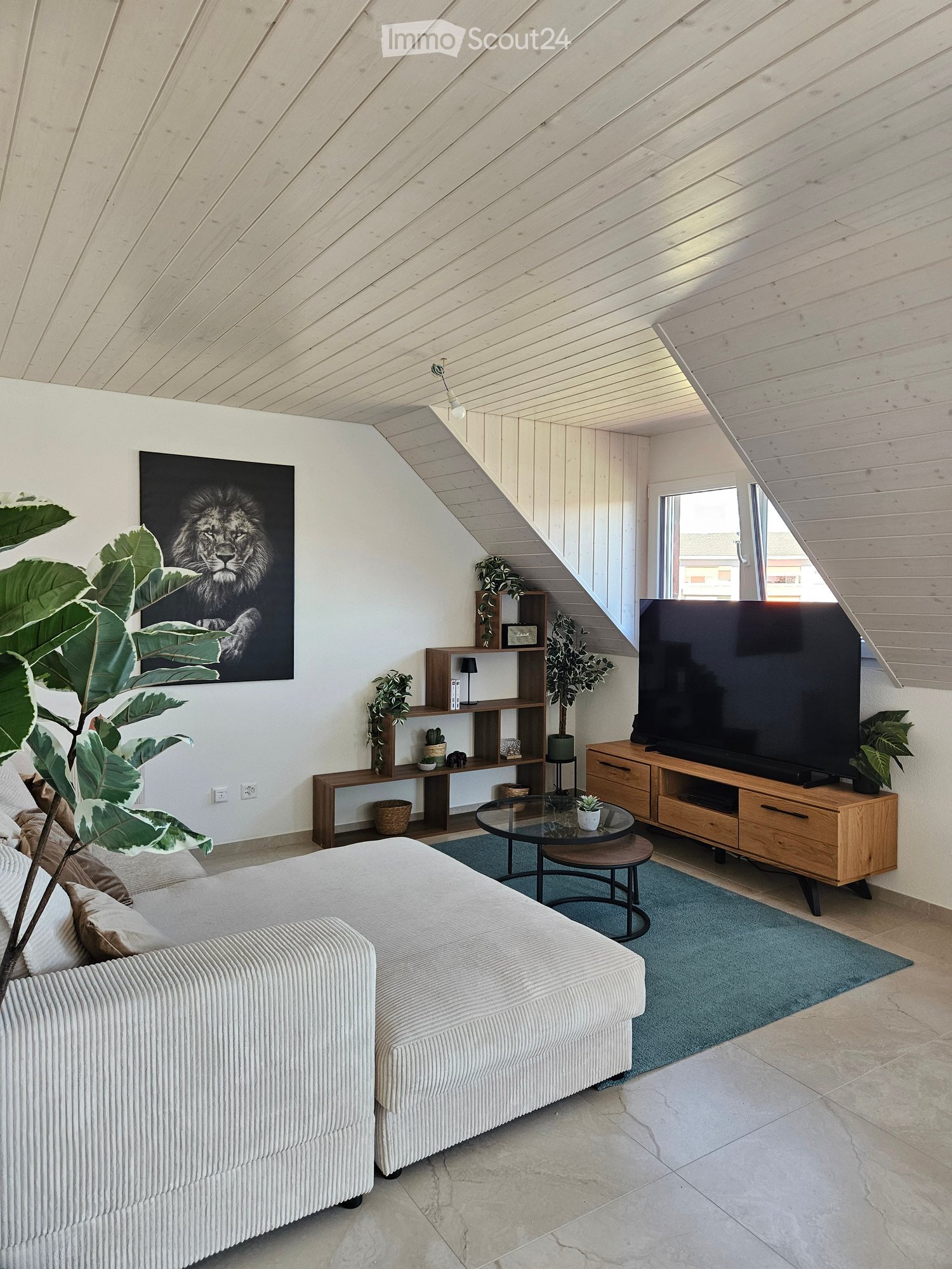 Modern living room with a white sofa, coffee table, TV stand, shelves, and potted plants. The room features a slanted wooden ceiling and large windows.