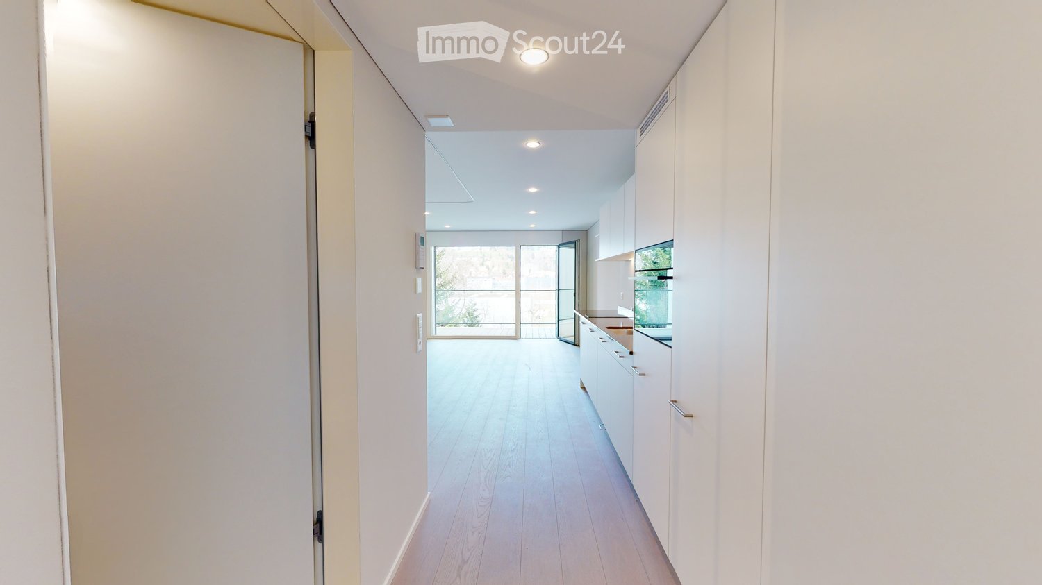 Bright, modern hallway with white walls, wooden flooring, recessed lighting, and a view into a kitchen area with white cabinets and a large window.