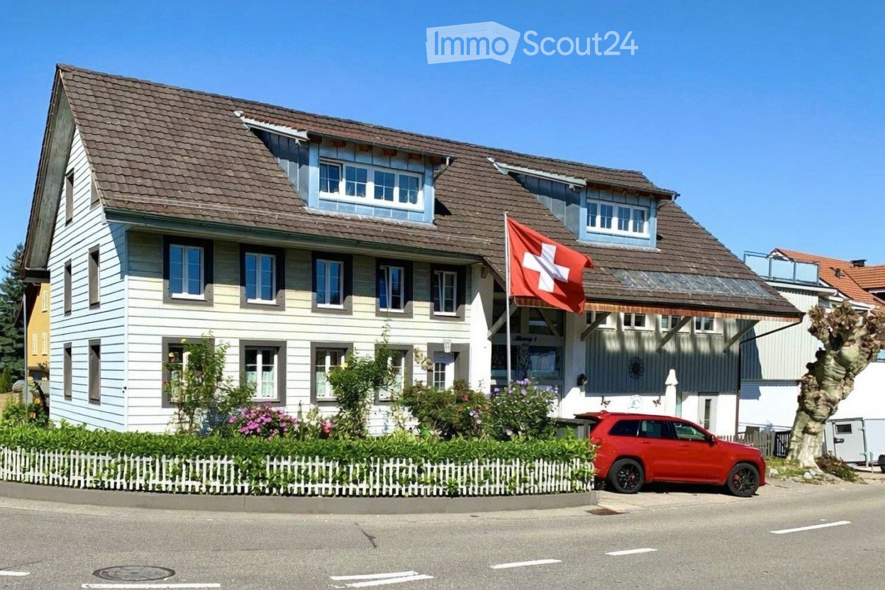 Two-story house with a brown tiled roof, white siding, multiple windows, and a Swiss flag. A red SUV is parked in front, and there is a small garden with a white picket fence.