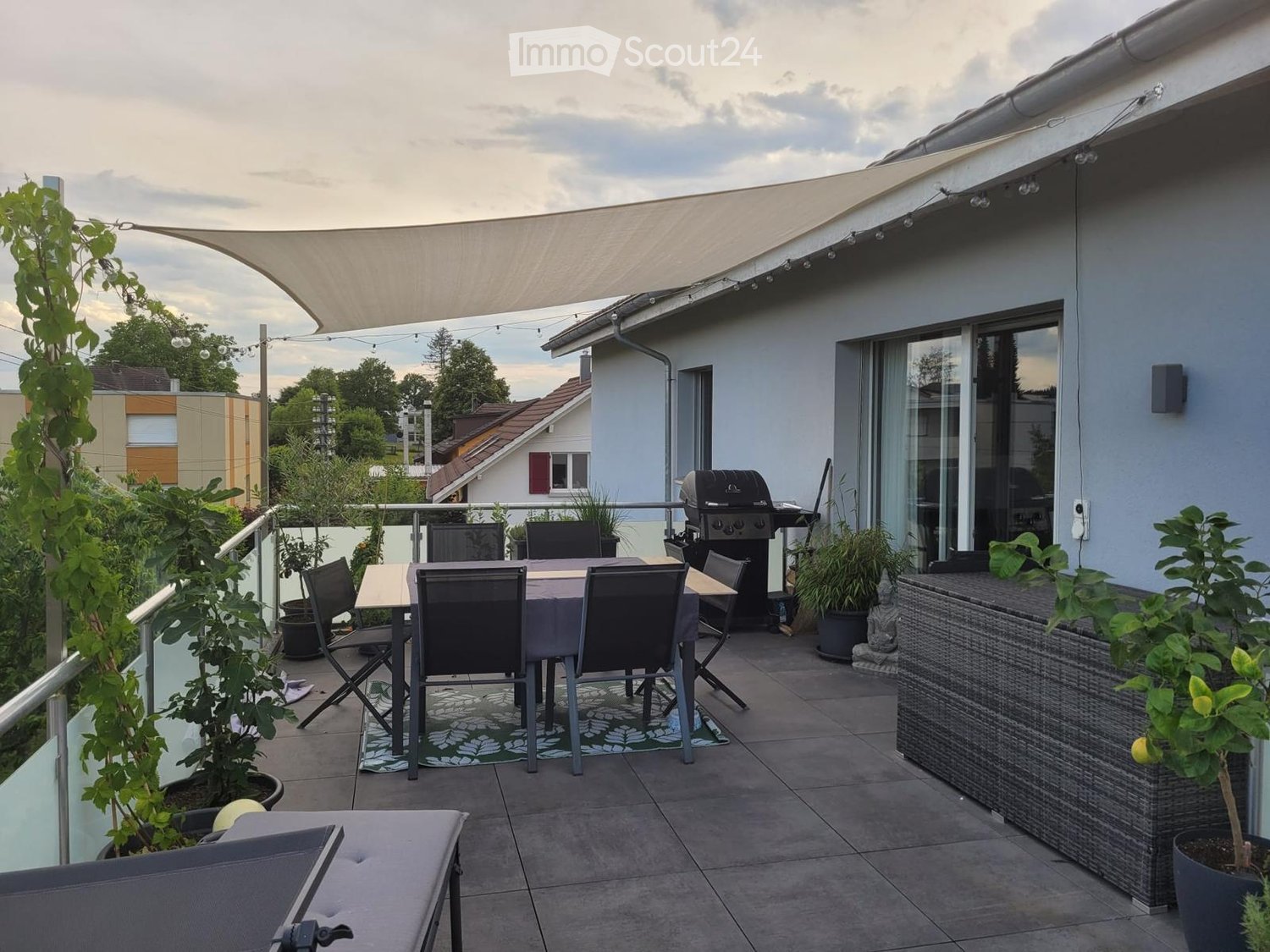 Balcony with a dining table, chairs, grill, storage box, and potted plants. Features a retractable awning and a view of the surrounding area.